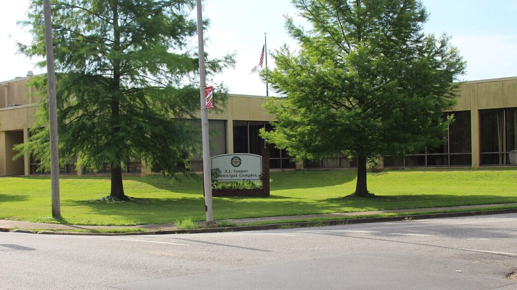 Government building with manicured lawn, flagpole, and signage at street corner
