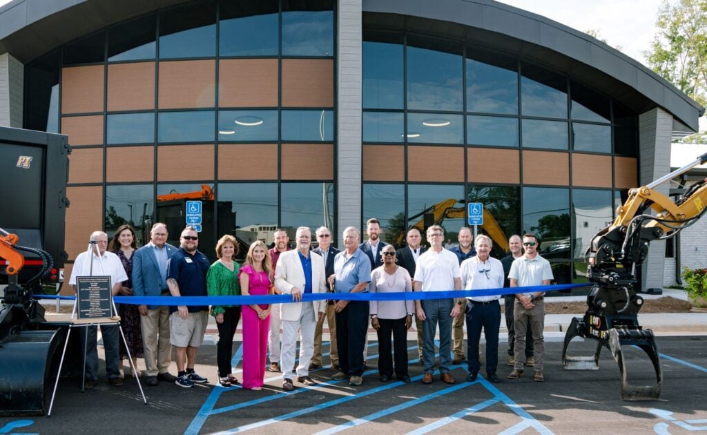 Group of officials and staff cutting blue ribbon at new public works facility with heavy equipment visible