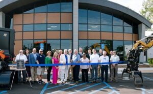 Group of officials and staff cutting blue ribbon at new public works facility with heavy equipment visible
