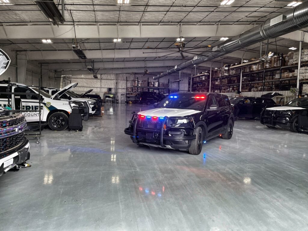 A police upfitting shop with a black Ford Police Interceptor Utility, its lightbar lit, alongside white sheriff SUVs under work