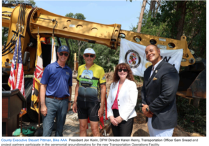 Four people stand in front of yellow construction equipment at a groundbreaking ceremony