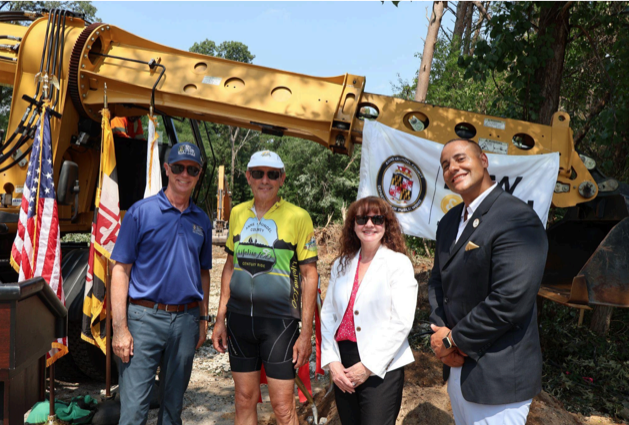 Four people stand in front of yellow construction equipment at a groundbreaking ceremony