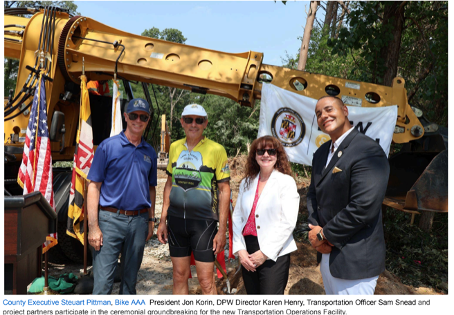 Four people stand in front of yellow construction equipment at a groundbreaking ceremony