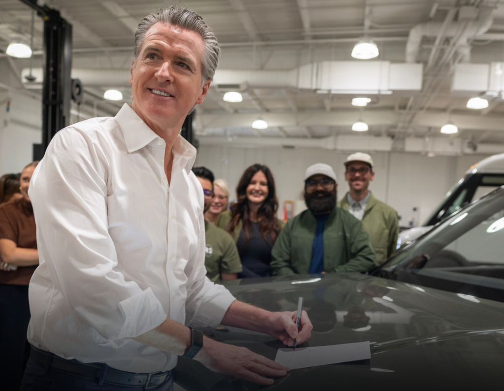 California Governor Gavin Newsom signing a document on the hood of an electric vehicle inside a service garage while staff look on