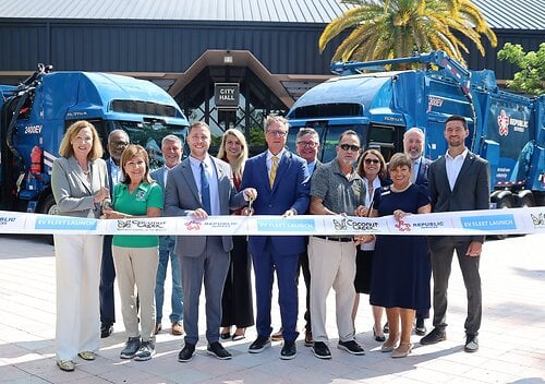 Coconut Creek officials holding an EV Fleet Launch ribbon in front of two blue electric refuse trucks outside City Hall