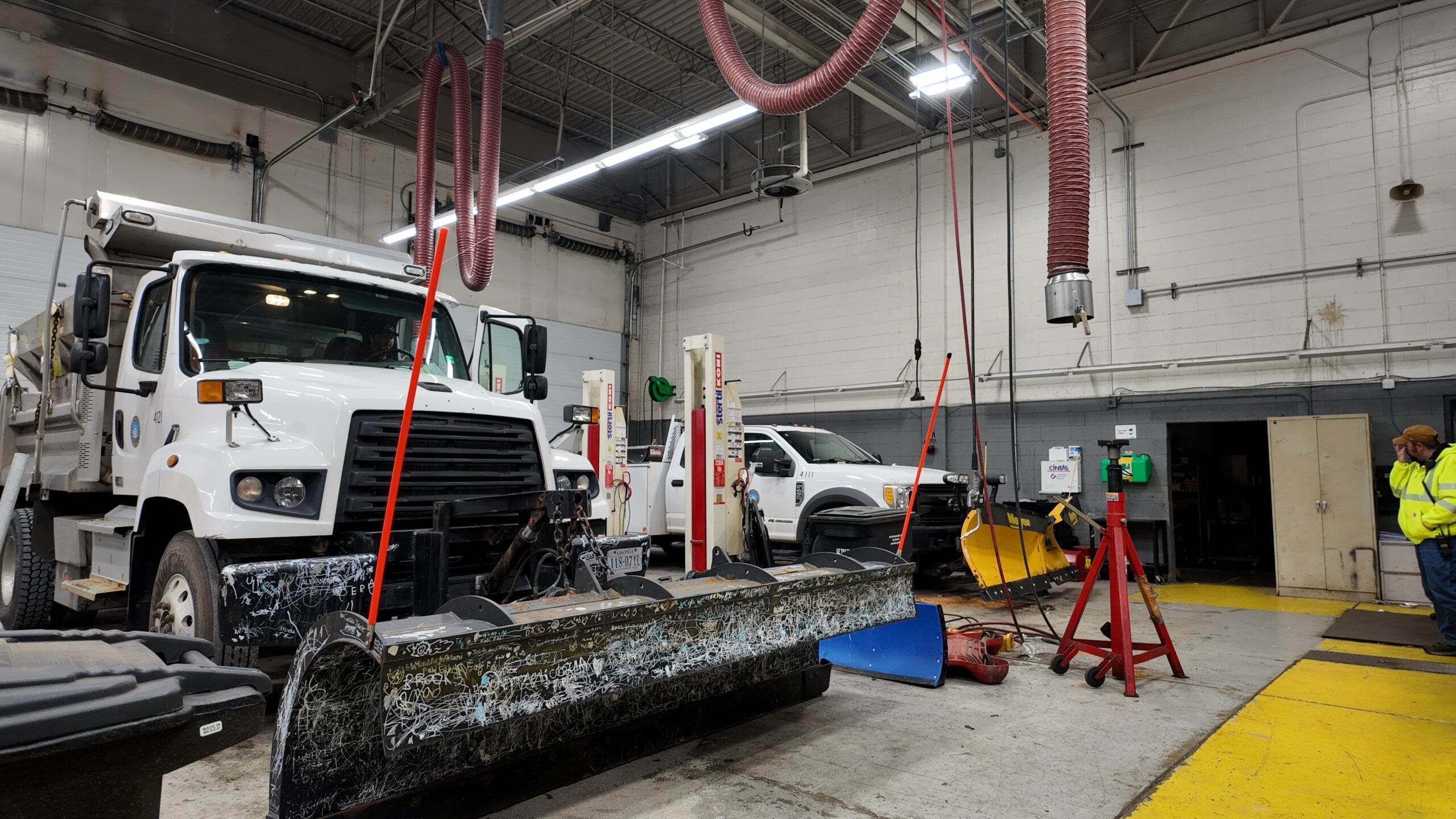 Municipal fleet maintenance shop with a plow-equipped dump truck, snow plow blades on the floor and overhead exhaust extraction hoses