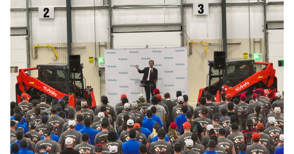 Man speaking at podium before large crowd in industrial warehouse with red Kubota equipment