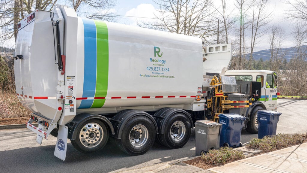 Recology automated side-loader garbage truck with blue and green stripe, parked curbside with collection bins
