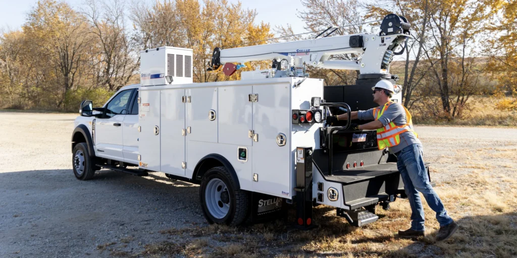 A technician in a high-visibility vest operating the controls of a white Stellar mechanic service truck with a crane, parked on gravel