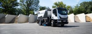 White and black street sweeper truck with rotating brush attachment parked in industrial yard