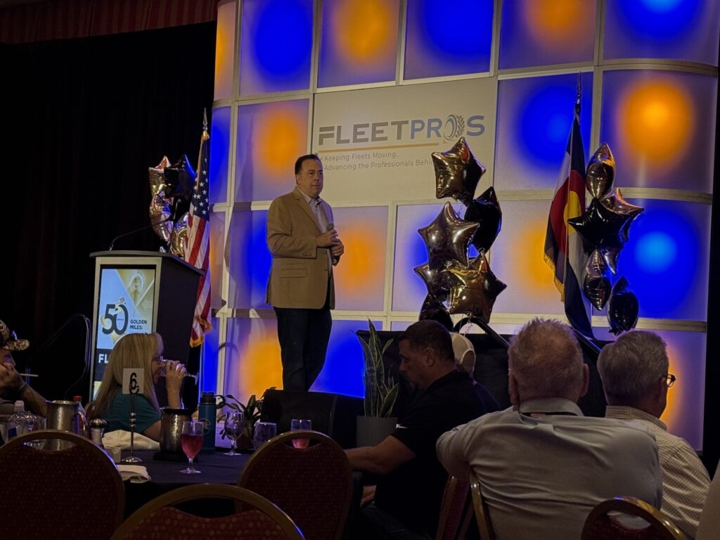 Man in tan blazer speaking at podium during FleetPros event with audience seated at tables