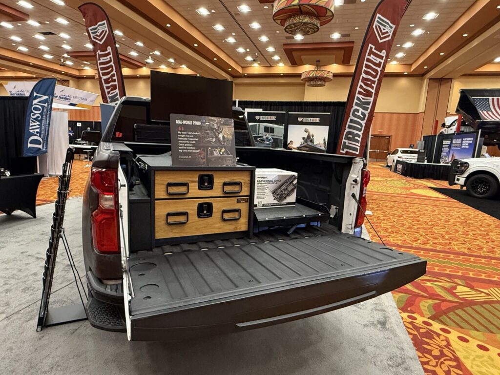 White pickup truck with open bed displaying wooden storage cabinetry and equipment at indoor trade show