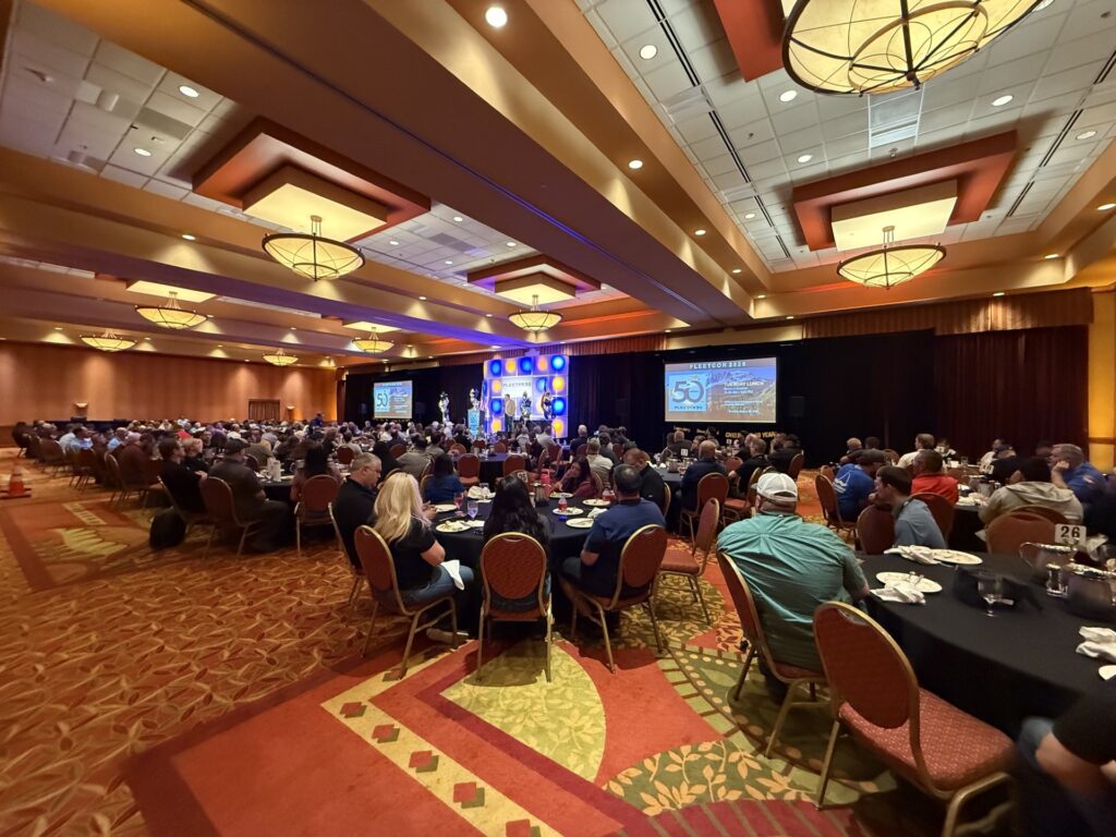 Large conference ballroom with hundreds of attendees seated at round tables facing stage with presentations and screens