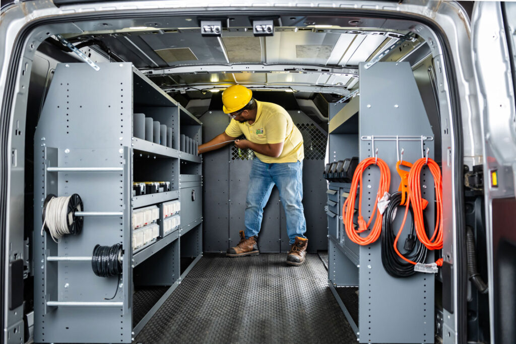Worker in yellow hard hat organizing shelving and coiled orange extension cords inside service van