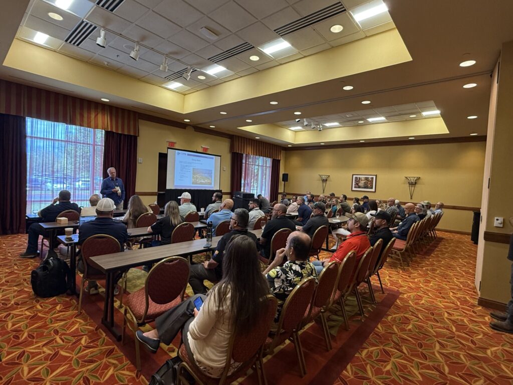 Large conference room with attendees seated at tables facing a presenter and projection screen