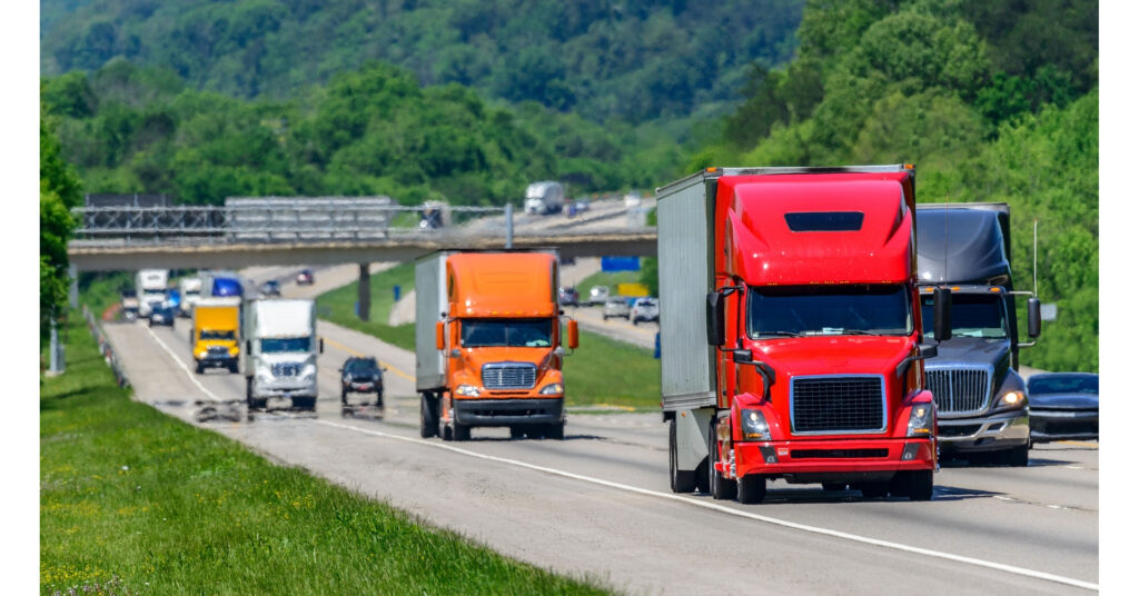 Multiple commercial trucks and vehicles traveling on multi-lane highway with overpass and forested hills