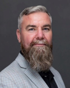 Professional headshot of a bearded man in light gray blazer and black shirt against gray background