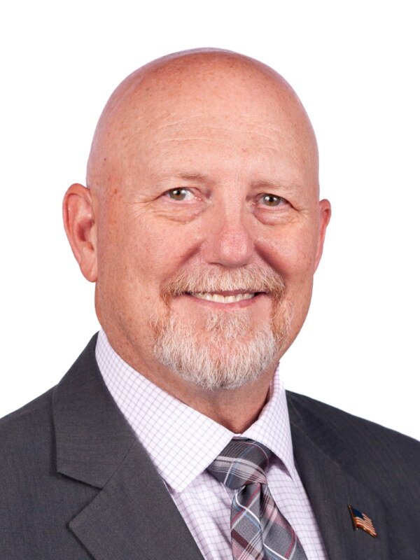Professional headshot of a bald man with a beard wearing a dark blazer, light purple checkered shirt, and plaid tie