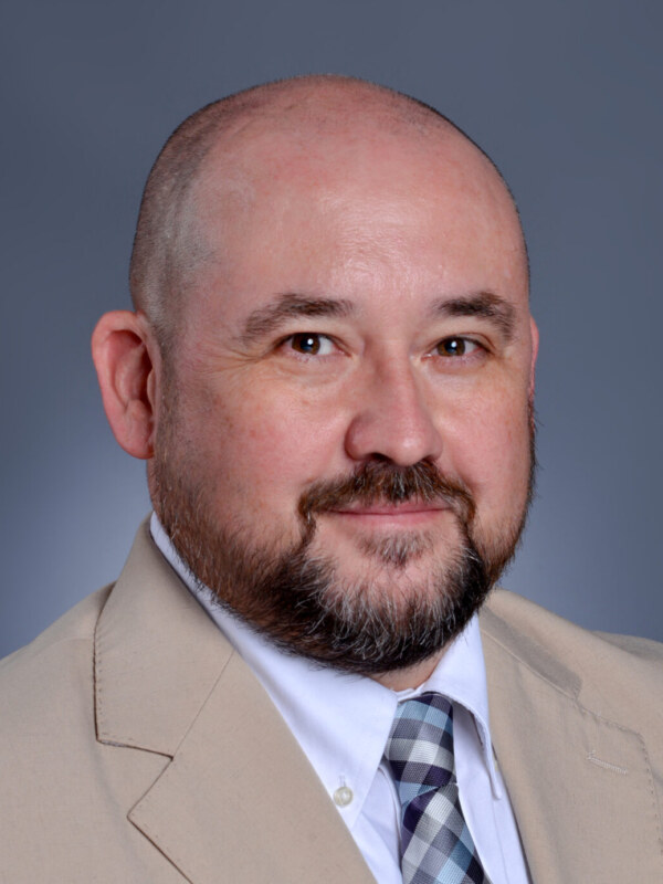 Professional headshot of a man in a beige blazer and patterned tie against blue background