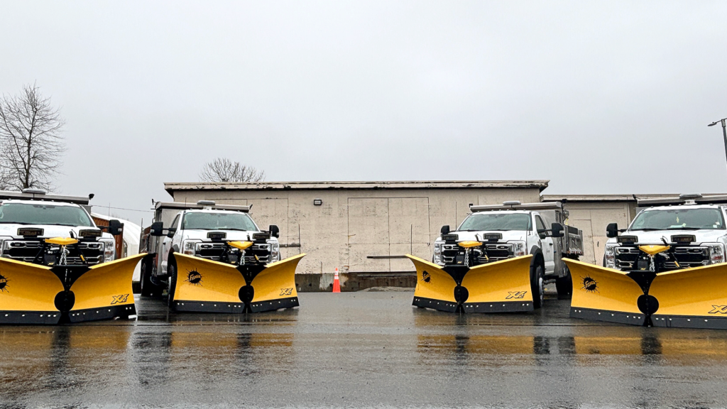 Four snow plow trucks with yellow plows lined up on wet pavement in front of a building