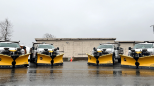 Four snow plow trucks with yellow plows lined up on wet pavement in front of a building