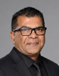 Professional headshot of man wearing black suit jacket, black dress shirt, and black-framed glasses against gray background
