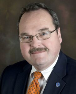 Professional headshot of man wearing glasses, dark suit, and orange patterned tie