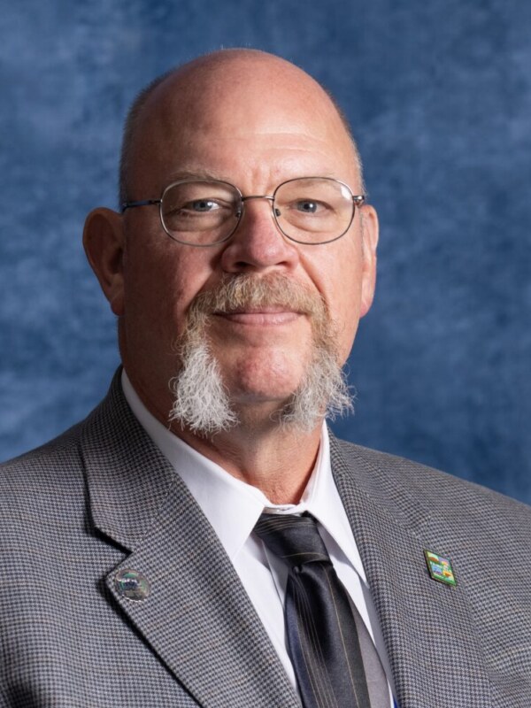 Professional headshot of bald man with beard wearing gray suit and dark tie against blue background