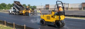 Yellow Sakai pneumatic tire roller compacting asphalt on parking lot with dump truck and crew members