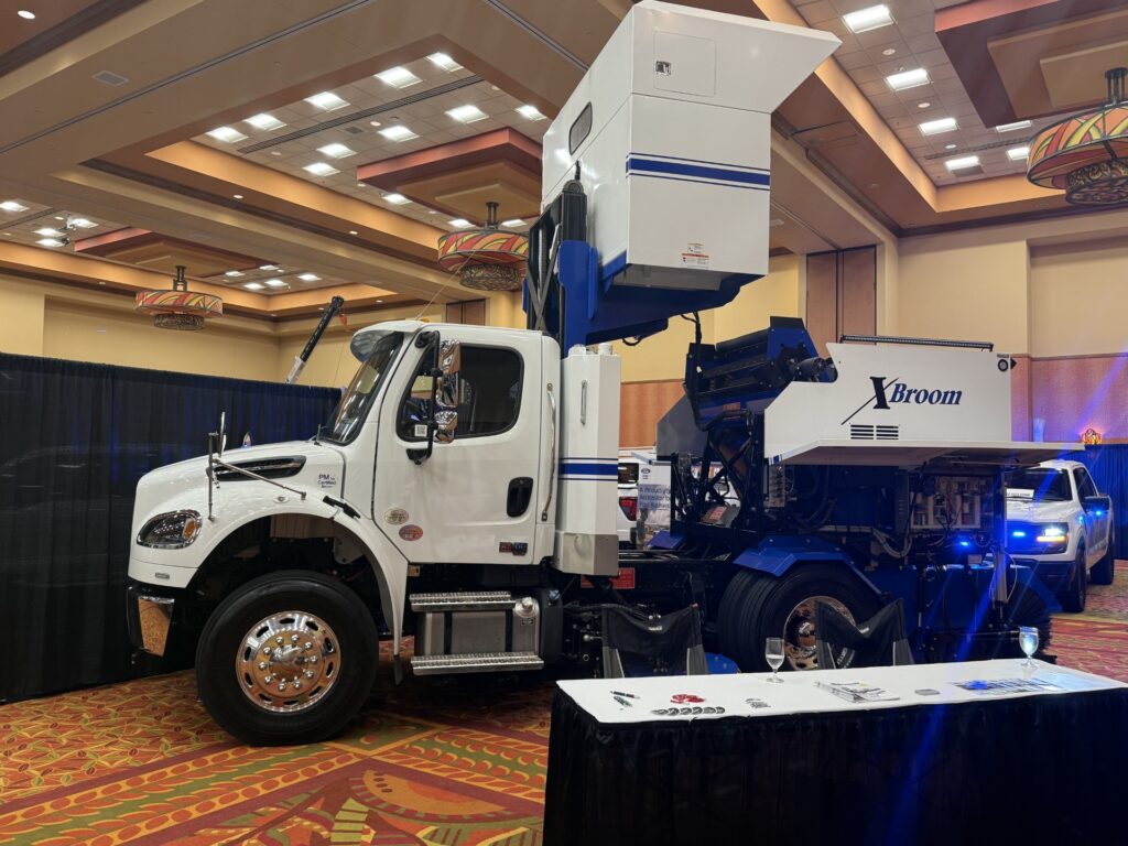 White street sweeper truck with blue Xbroom branding displayed indoors at conference