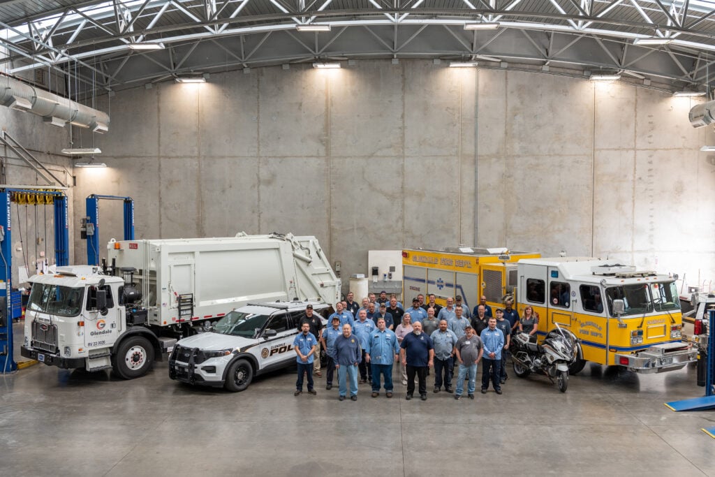 Team of municipal workers posing with garbage truck, police vehicle, and fire truck indoors