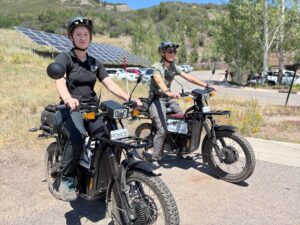 Two police officers on electric bicycles wearing helmets, with solar panels and parked vehicles visible in background