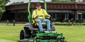 Operator in high-visibility vest on green zero-turn riding mower on manicured lawn