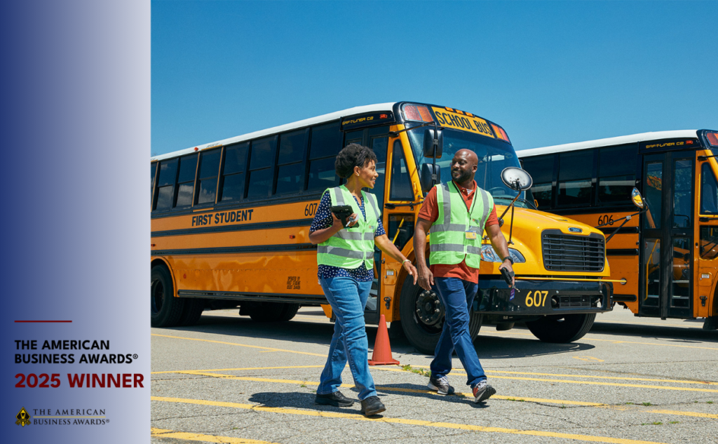 Two safety personnel in high-visibility vests stand between yellow school buses under clear sky