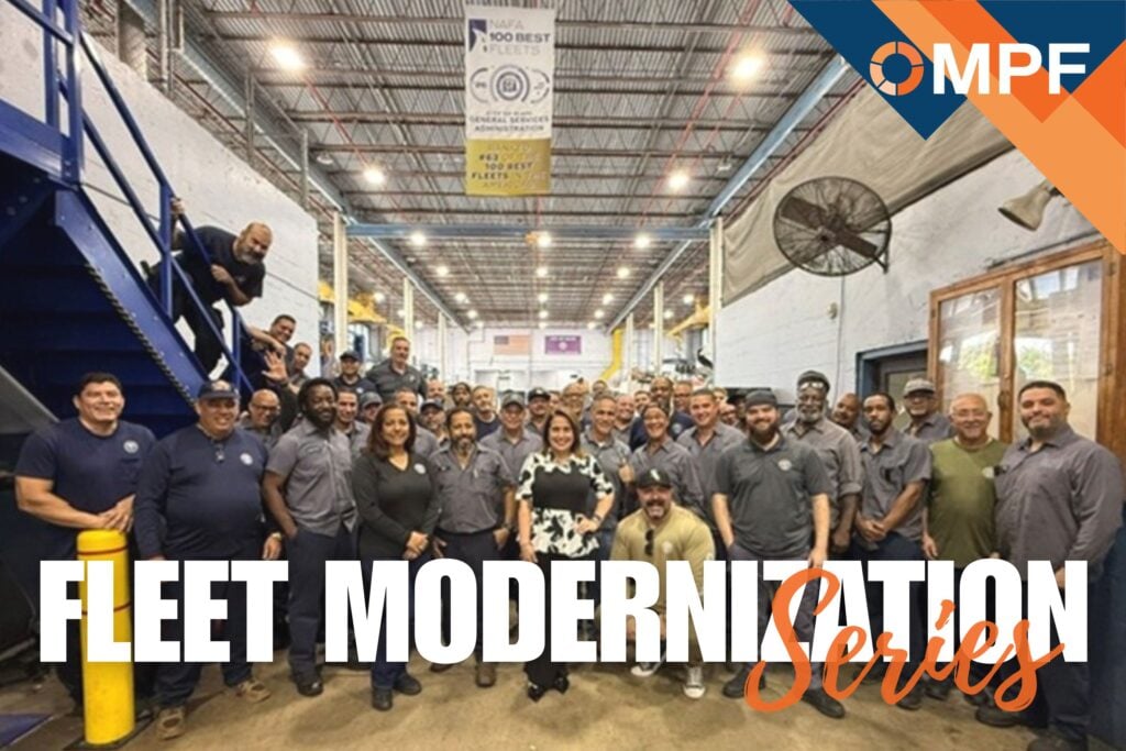 Large group of fleet workers posing together in industrial warehouse facility with OMPF logo visible