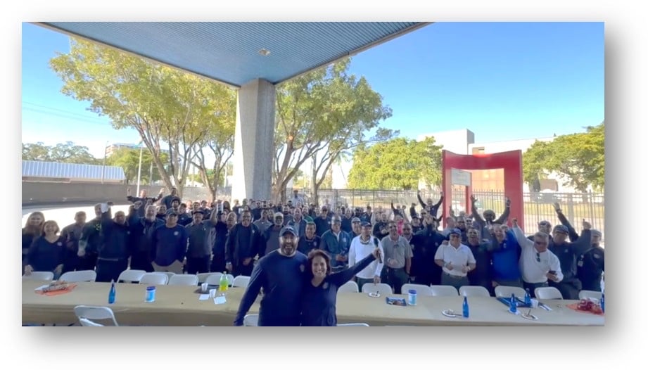 Large group of people gathered under covered outdoor structure with trees in background