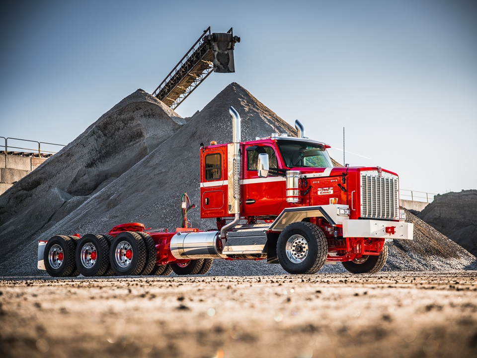 Red heavy-duty truck with aggregate conveyor system and material stockpiles at industrial site
