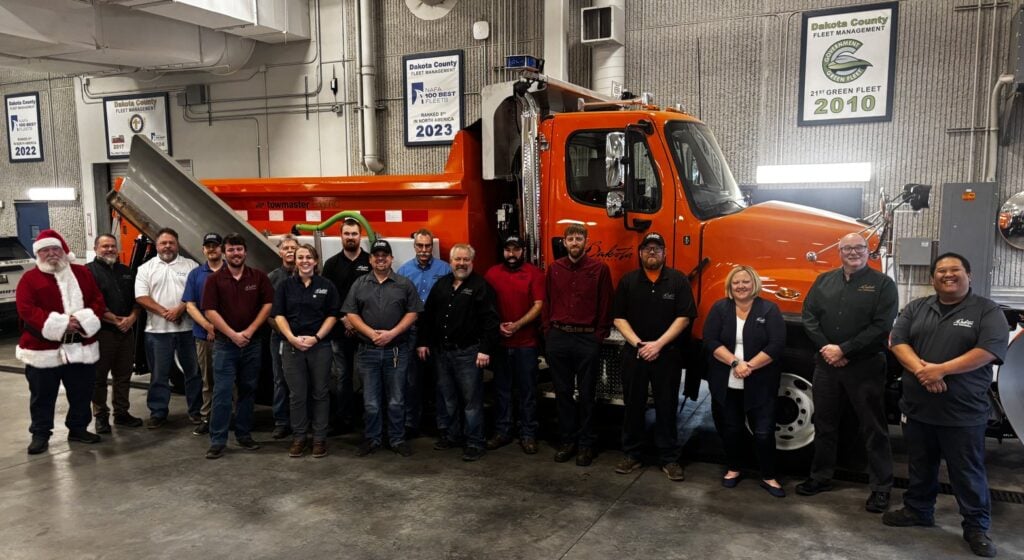 The Dakota County Fleet Management staff, joined by Santa Claus, stand in front of an orange county plow truck in the shop beneath the fleet's 100 Best Fleets and Green Fleet award banners.