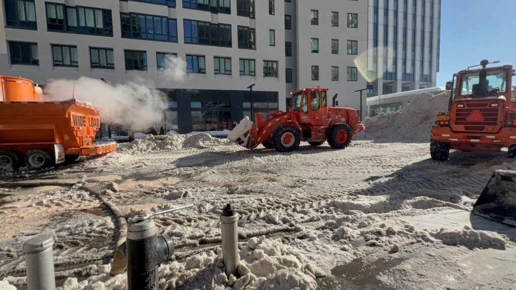 Department of Sanitation loaders move piled snow beside steaming snow melters in front of apartment buildings