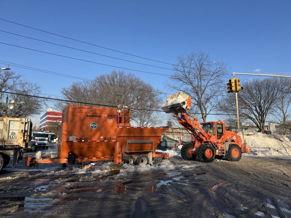 An orange Department of Sanitation front-end loader lifts snow into a trailer-mounted snow melter on a Brooklyn street