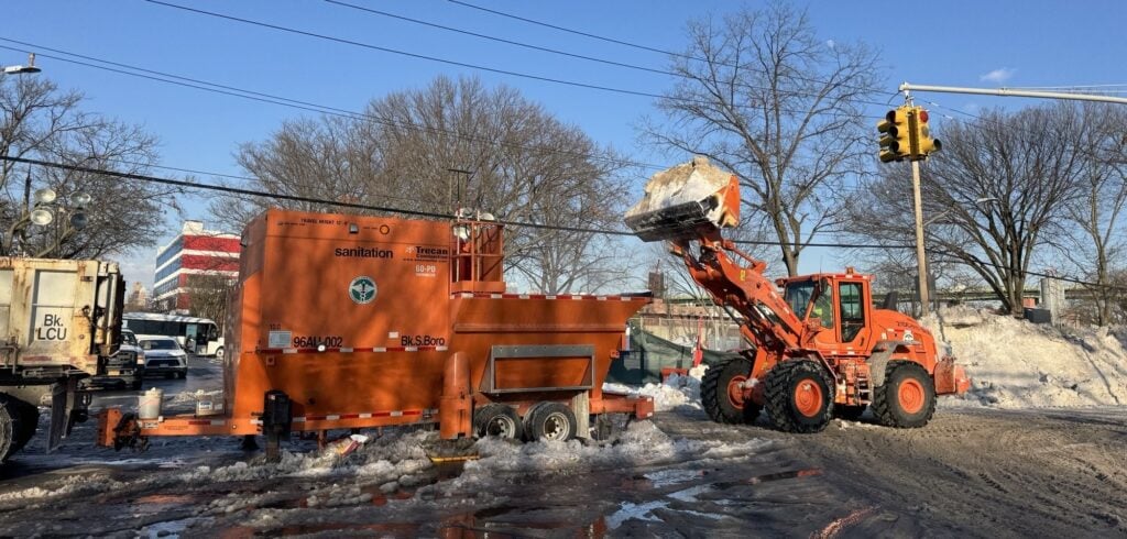 An orange Department of Sanitation front-end loader lifts snow into a trailer-mounted snow melter on a Brooklyn street