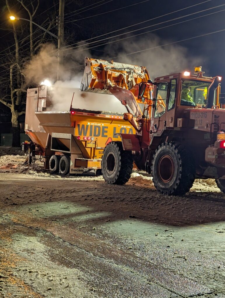 A loader dumps snow into a steaming snow melter on a Queens street at night