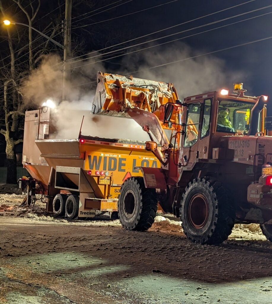 A loader dumps snow into a steaming snow melter on a Queens street at night