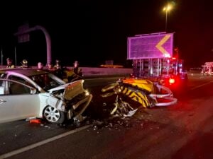 Nighttime highway collision scene with damaged white sedan, yellow vehicle, emergency responders, and illuminated truck with pink signage