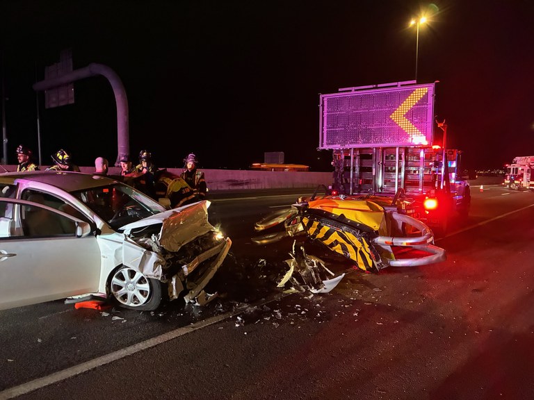 Nighttime highway collision scene with damaged white sedan, yellow vehicle, emergency responders, and illuminated truck with pink signage