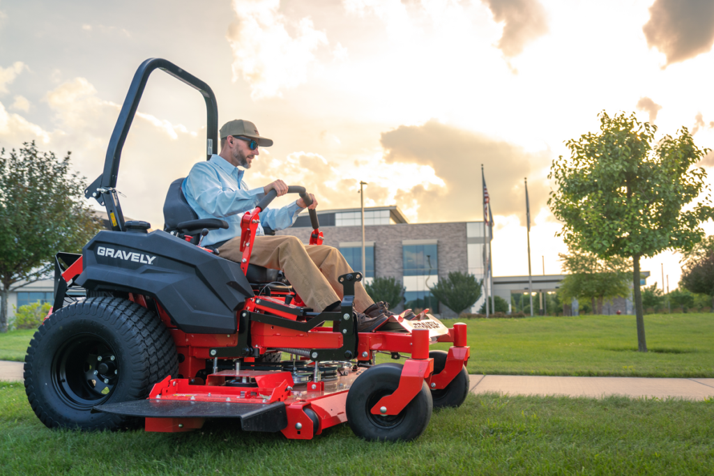 Operator on red Gravely zero-turn mower in manicured park setting with building in background