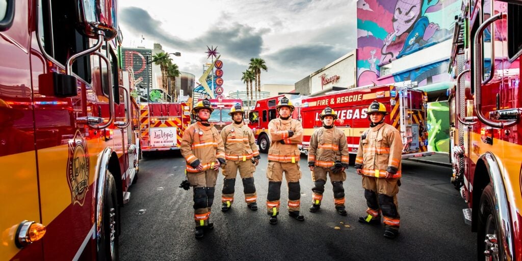 Six firefighters in tan uniforms standing between red and yellow fire trucks