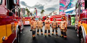 Six firefighters in tan uniforms standing between red and yellow fire trucks