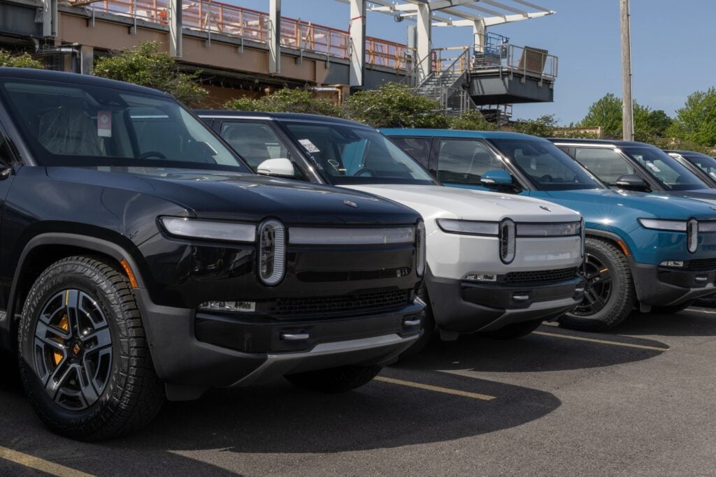 Row of four electric SUVs parked in lot with industrial infrastructure behind
