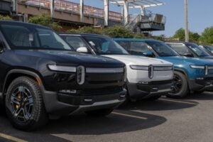 Row of four electric SUVs parked in lot with industrial infrastructure behind